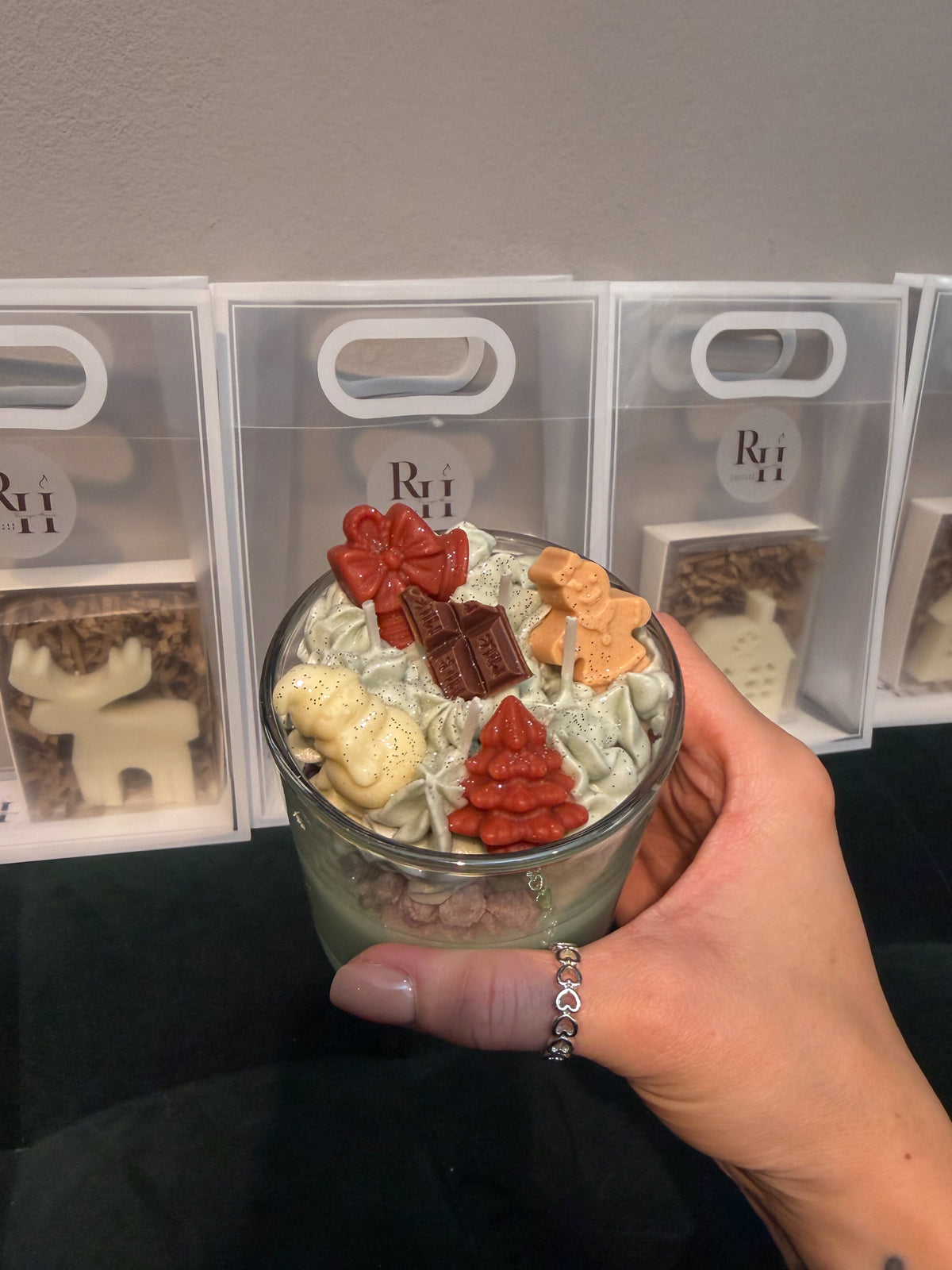 Hand holding a handmade green Christmas dessert candle decorated with whipped wax, gingerbread figures, a Christmas tree and chocolate-shaped wax decorations, with small Christmas gifts for workshop participants in the background.