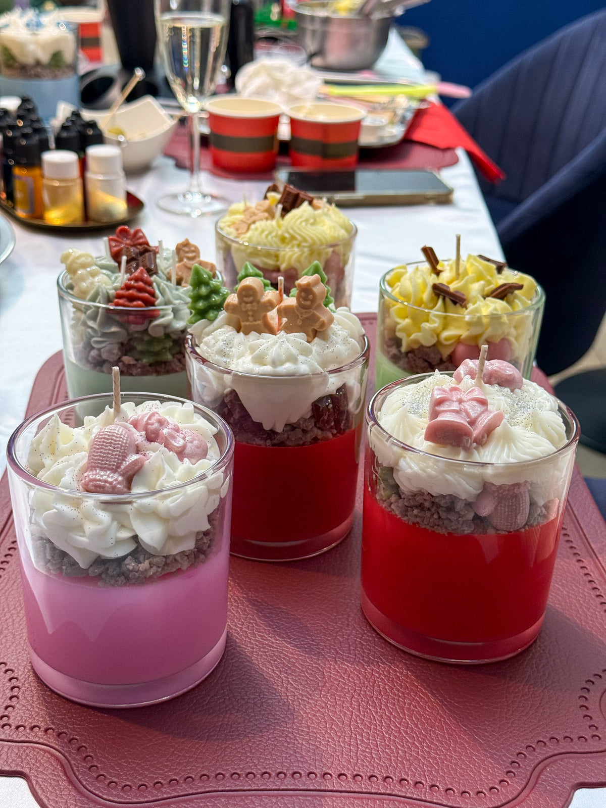 Six handmade Christmas dessert candles in glass cups, decorated with whipped wax, gingerbread figures, Christmas trees and festive decorations at a candle-making workshop in Vienna.