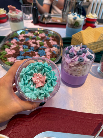 Hand holding a handmade Christmas dessert candle with turquoise whipped wax and a pink bow decoration, next to a purple candle topped with wax blueberries at a candle-making workshop in Vienna.