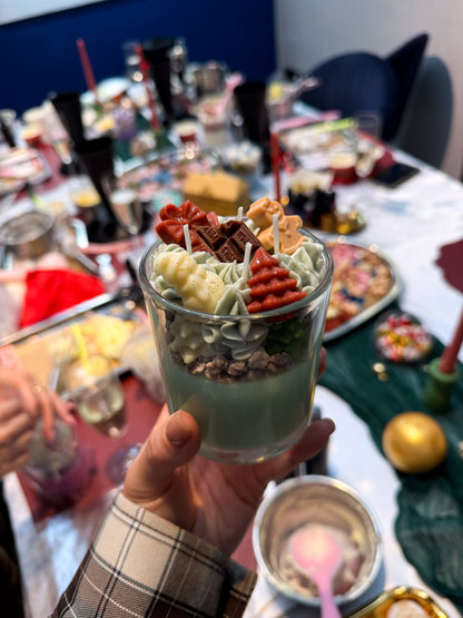 Hand holding a handmade green Christmas dessert candle in a glass, decorated with whipped wax, Christmas trees, gingerbread figures and chocolate-shaped wax decorations at a candle-making workshop in Vienna.