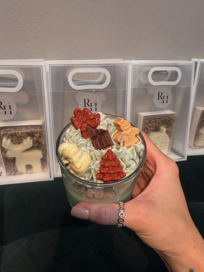 Hand holding a handmade green Christmas dessert candle decorated with whipped wax, gingerbread figures, a Christmas tree and chocolate-shaped wax decorations, with small Christmas gifts for workshop participants in the background.