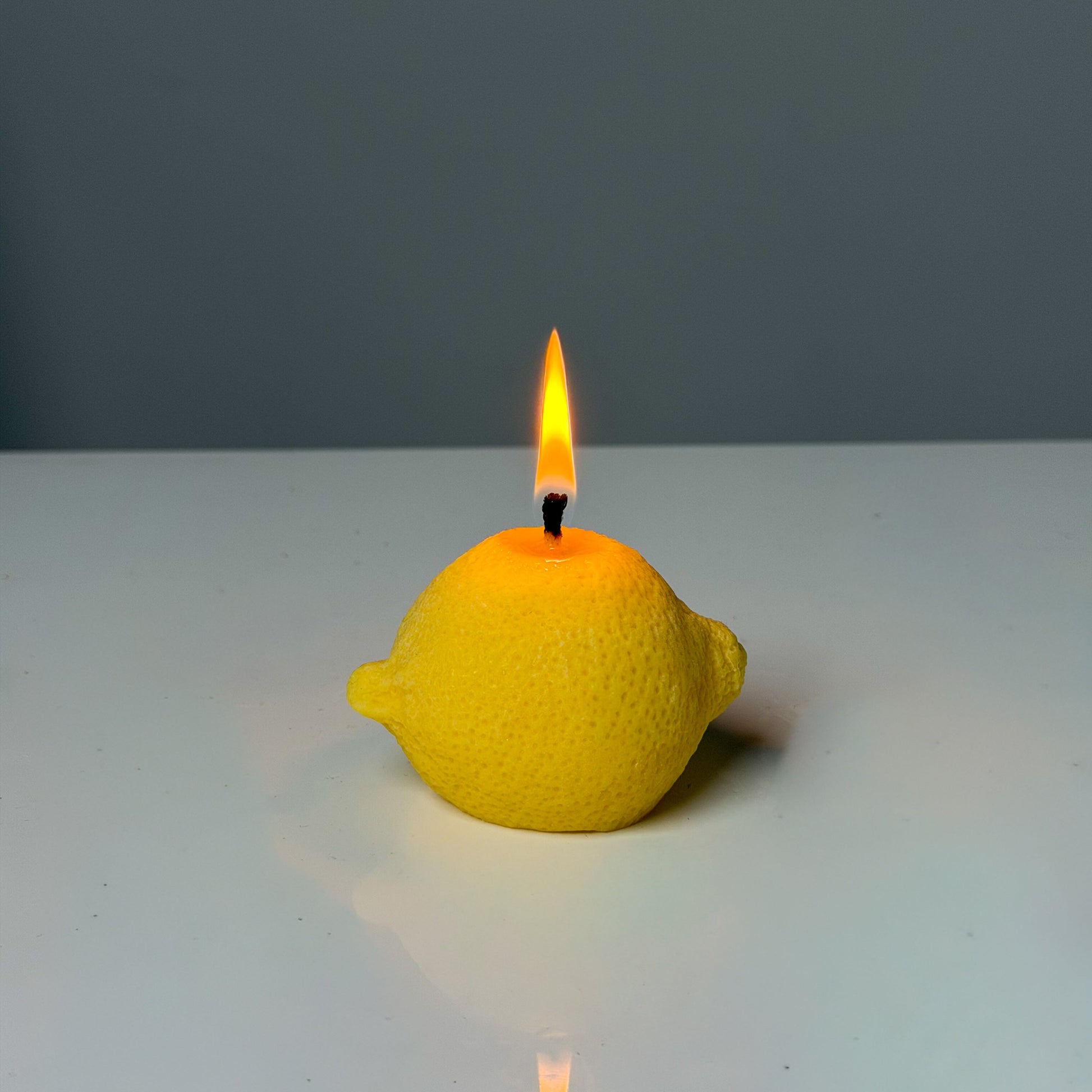 Yellow lemon-shaped candle with a lit wick on a white surface and gray background