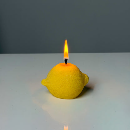 Yellow lemon-shaped candle with a lit wick on a white surface and gray background