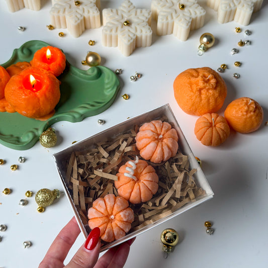 Hand holding a gift box of three mandarin candles with paper filling among Christmas decorations.
